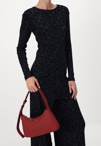 Rode handtas gemaakt van glad materiaal met een gebogen vorm, vastgehouden door een persoon die een langemouwen, donker patroon top en losse broeken draagt.