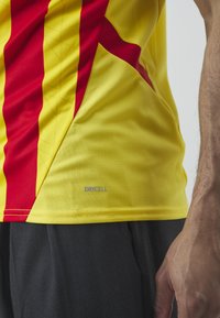 Gul atletisk trøje med røde striber; letvægts, tekstureret stof; har "DRYCELL" logo nær nederkanten; tætsiddende design.