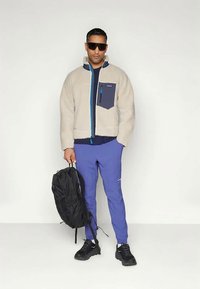 Μπουφάν fleece σε μπεζ με ναυτικό τσέπη, πράσινο φερμουάρ και μαύρο παντελόνι. Μαύρο σακίδιο πλάτης και γυαλιά ηλίου συμπληρώνουν την εμφάνιση.