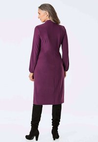 Langärmliges, lila Kleid aus glattem Stoff, mit gerader Schnittführung und rückseitiger Nahtdetails. Kombiniert mit kniehohen schwarzen Stiefeln.