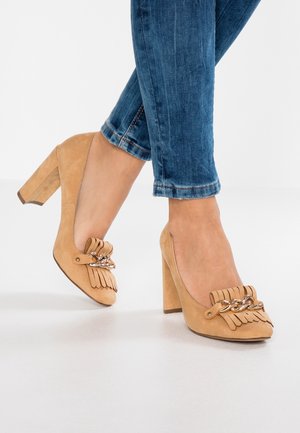 Béžové semišové lodičky na vysokém podpatku s třásněmi a řetízkem. Čtvercové podpatky a zaoblené špičky, kombinované s modrými džíny.