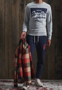 Sudadera gris con logotipo azul marino, pantalones de chándal oscuros, zapatillas blancas y una camisa de franela de cuadros rojos con cuadrados verdes y beige.