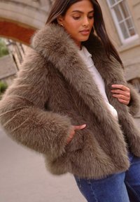 Kailiniai švarkas iš dirbtinio pūko taupe spalva su plačiu apykakle ir švelnia tekstūra. Pasižymi šoniniais kišenėmis ir nukritusiais petimis, derinamas su baltu marškinėliu ir džinsais.