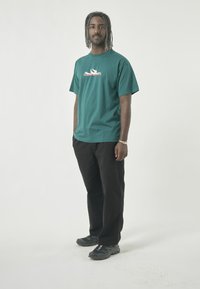 Green cotton t-shirt with a graphic print and short sleeves, paired with black pants and grey athletic shoes, standing against a white background.