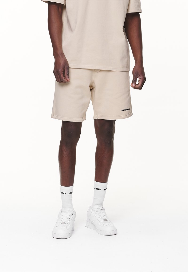 Homme portant un short beige et un t-shirt avec le logo « Pegador », des chaussettes blanches et des baskets blanches, debout devant un fond blanc.