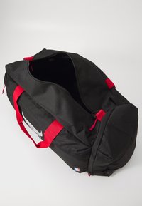 Bolsa de deporte negra con asas rojas, cremallera parcialmente abierta y pequeño logo de la NBA sobre fondo blanco.