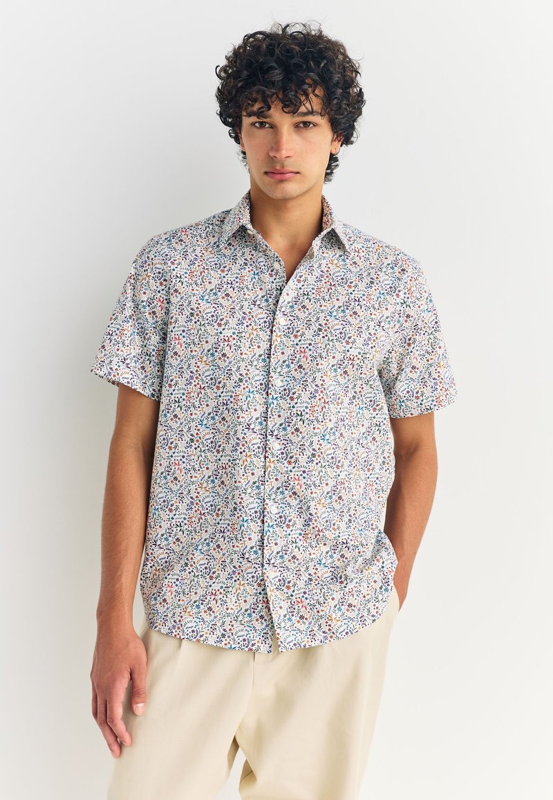 Chemise à manches courtes avec boutons, présentant un motif floral multicolore sur fond blanc, confectionnée dans un tissu léger, avec une coupe décontractée.