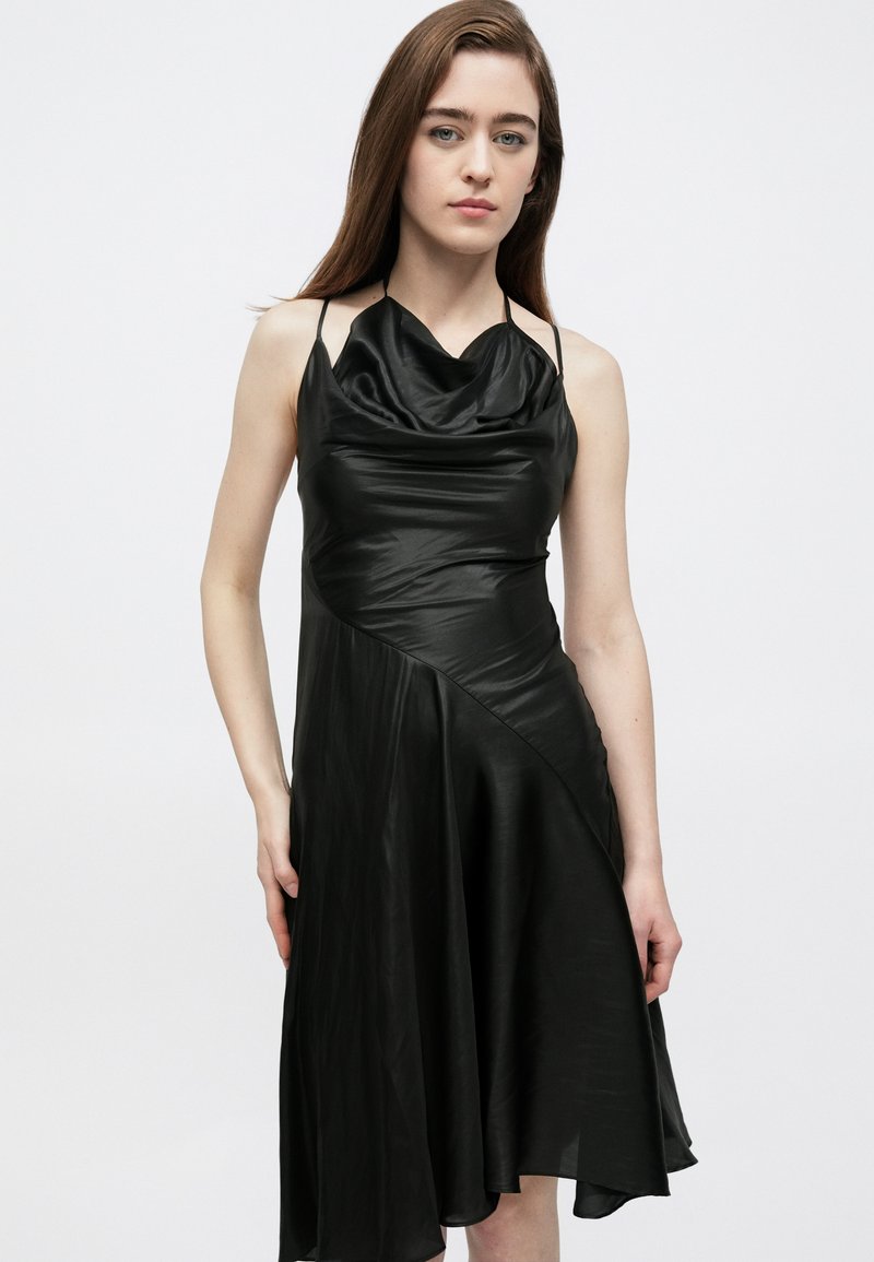 Sort kjole med halstørklædehals og tynde stropper, lavet af et glat materiale. Har en diagonal søm og et flydende skørt, der falder til midt på læggen.