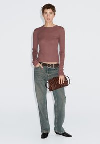 Langärmliges bordeauxrotes Oberteil, blaue weite Jeans und schwarze spitze Schuhe. Hält eine dunkelbraune Leder-Clutch mit kurzen Griffen.