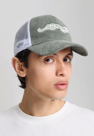 Młody mężczyzna noszący zieloną sztruksową i białą siateczkową czapkę Ed Hardy oraz gładką białą koszulę na jednolitym tle.