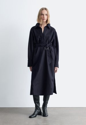 Navy blue shirt dress with long sleeves, belted waist featuring a silver buckle, and side slits; worn with black knee-high leather boots.