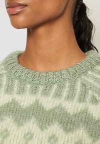 Jasnozielony sweter z dzianiny z teksturowanym wzorem i kremowymi akcentami. Żebrowany kołnierz dodaje detalu luźnemu dekoltowi.