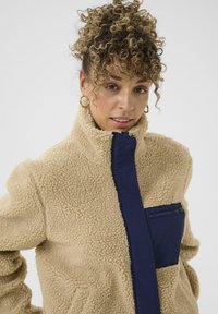 Veste en polaire beige avec un col haut et une poche zippée bleu marine. Elle possède une finition texturée et un panneau en tissu contrasté.