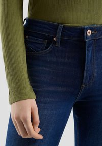 Jeansy z ciemnoniebieskiego denimu o wysokim stanie, z miedzianymi guzikami i gładką fakturą, zestawione z teksturowanym oliwkowo-zielonym topem.