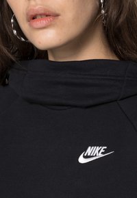 Svart sweatshirt med hög krage och vit Nike-logga. Har ett mjukt, texturerat tyg och kontrasterande sömmar på axlarna.
