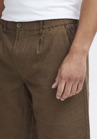 Barna, könnyű nadrág, pleátolt elülső résszel, rugalmas derékpánttal és gombos záródással. Az anyagnak sima felülete van, finom fénylással.