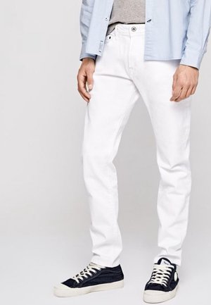 Homme portant un pantalon blanc, des baskets noires et blanches, une chemise bleu clair et un sous-vêtement gris, debout devant un fond uni.