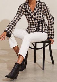 Musta-valkoinen houndstooth-kuvioinen bleiseri, jossa on vyötärönauha ja leveät hihat, yhdistettynä valkoisiin slim-fit-housuihin ja mustiin nilkkureihin.