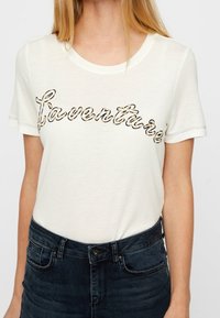 Mujer con una camiseta blanca de manga corta con la palabra "fourthree" en un diseño de leopardo en el pecho, acompañada de jeans de denim oscuros.