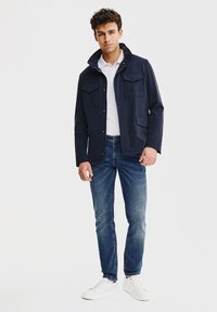 Granatowa wodoodporna kurtka z kołnierzem, z dwiema kieszeniami na piersi, noszona na białej koszulce polo i niebieskich dżinsach, w połączeniu z białymi sneakersami.