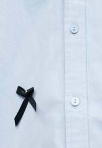 Ανοιχτό μπλε πουκάμισο με κουμπιά και μικρή μαύρη κορδέλα ευαισθητοποίησης καρφιτσωμένη κοντά στα κουμπιά στην αριστερή πλευρά.