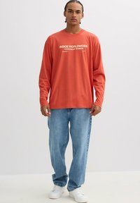 Camiseta de manga larga color óxido con texto gráfico blanco, combinada con jeans de corte holgado en azul claro y zapatillas blancas, de pie sobre un fondo neutro.