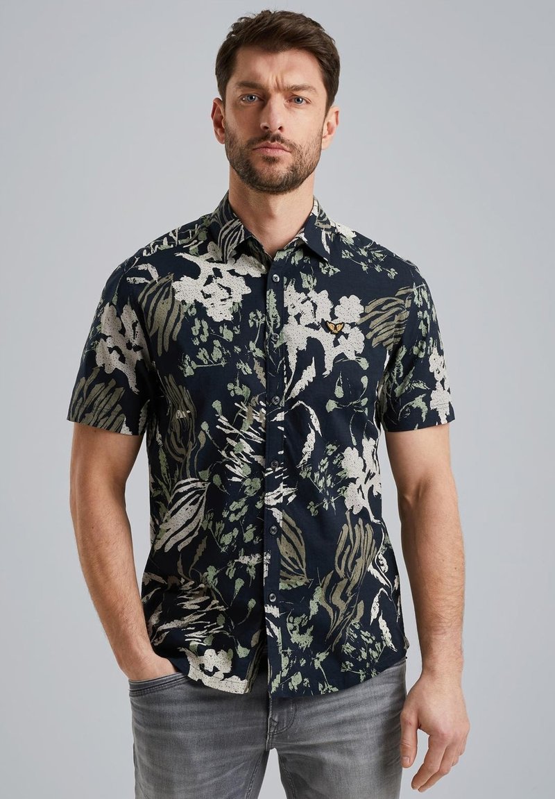 Camisa de manga corta en azul marino con estampado floral en blanco y verde; cuenta con un cuello abotonado y un pequeño logo en el pecho. Hecha de algodón.