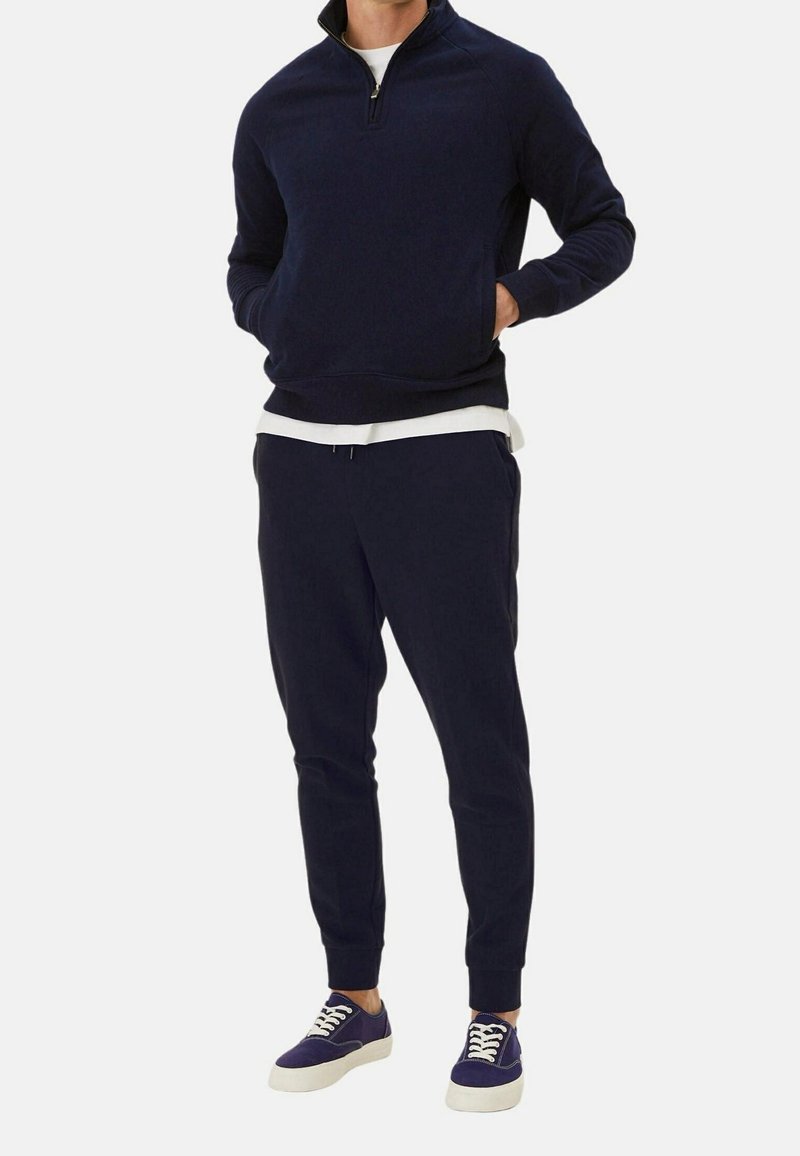 Homme portant un sweat-shirt bleu marine à fermeture éclair quart, un pantalon de jogging assorti, un T-shirt blanc en dessous et des baskets bleues avec des lacets blancs.