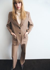 Woman with long blonde hair wearing a beige pantsuit and cream turtleneck, standing with hands in pockets against white wall.