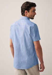 Šviesiai mėlyna trumpomis rankovėmis marškiniai, pagaminti iš struktūrinės medžiagos, su mygtukų užsegimu ant apykaklės ir kreivu apačios kraštu. Atvirkštinis vaizdas rodo siūles ir siuvimą.