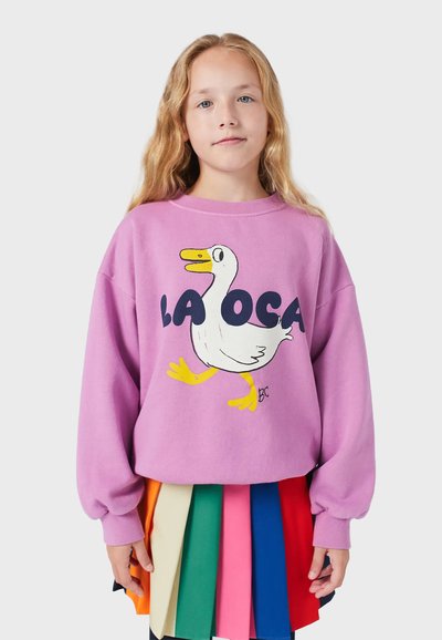 Fialová flísová mikina s grafikou kresleného kačera a textom "LA OCA", oblečená s viacfarebnou plisovanou sukňou.