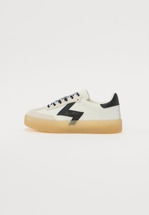 Sneakers with a creamy white leather upper, black lightning bolt accent, patterned black laces, and a semi-transparent gum sole.