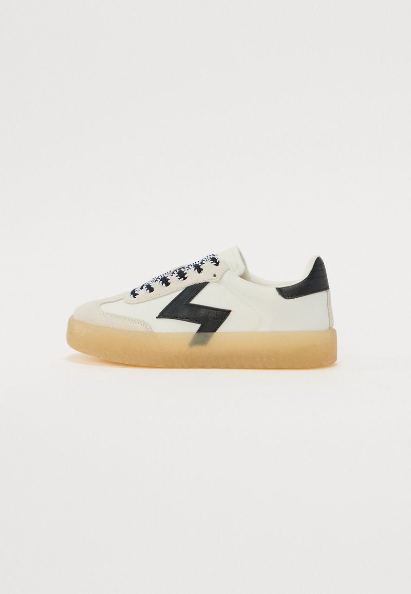 Sneakers med en kremhvit skinnøvre, svart lynboltakkent, mønstrede svarte snøringer og en delvis gjennomsiktig gummisåle.