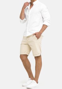 Biała lniana koszula z guzikami i podwiniętymi rękawami, zestawiona z beżowymi szortami z odwiniętymi brzegami oraz białymi trampkami.