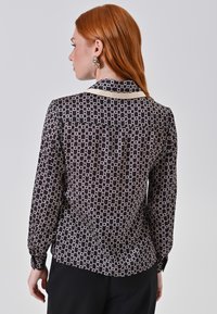 Donna dai capelli rossi che indossa una blusa nera a maniche lunghe con motivi a catena, abbinata a pantaloni neri, vista di schiena su uno sfondo grigio.