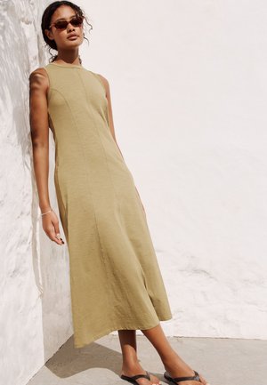 Mujer con vestido midi oliva sin mangas y gafas de sol se apoya contra una pared blanca texturizada, de pie sobre un suelo de cemento iluminado por el sol con sandalias.