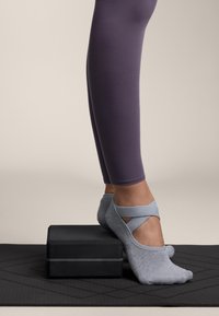 Grå låga strumpor med ett korsad remdesign på en svart yogablock, i kombination med lila åtsittande leggings på en texturerad matta.