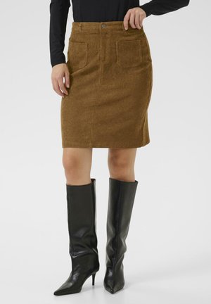 Jupe en velours côtelé marron avec une silhouette ajustée et deux poches arrière, accompagnée de bottes en cuir noires à talons pointus montant aux genoux.