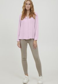 Blusa rosa chiaro a maniche lunghe con colletto, abbinata a pantaloni beige slim e sneakers bianche. Tessuto morbido con una vestibilità rilassata.
