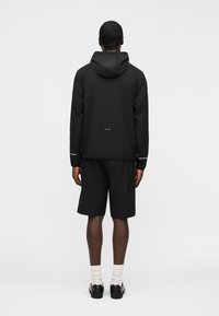 Άνδρας που φοράει μαύρο μπουφάν με κουκούλα και ταιριαστά σορτς στέκεται απέναντι σε λευκό τοίχο, φοράει λευκές κάλτσες και μαύρα παπούτσια.