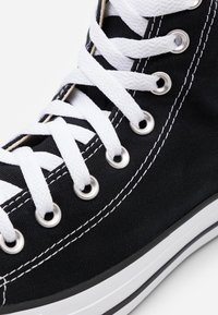 Czarne sneakersy typu high-top na czarnej podeszwie, z białymi sznurówkami, srebrnymi oczkami i kontrastującymi białymi szwami. Guma podeszwy z czarno-białymi paskami.