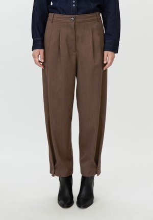 Woman wearing high-waist brown pleated trousers with buttoned cuffs and black ankle boots, paired with a dark denim shirt.