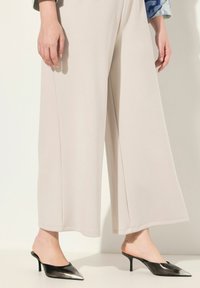 Pantalons larges beiges en tissu lisse avec une finition soignée, associés à des mules noires à talons aigus au design élégant.
