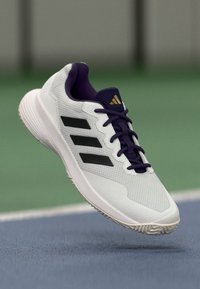 Weißer Sportschuh mit schwarzen Streifen, lila Akzenten, strukturiertem Obermaterial und Gummisohle. Entwickelt für die Leistung bei Sportaktivitäten.