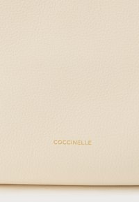 Superficie in pelle di colore crema con texture, con il piccolo marchio COCCINELLE impresso in oro al centro, vicino al bordo inferiore.