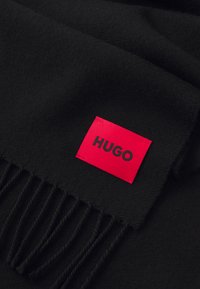 Sort tørklæde lavet af blødt materiale, med frynser i den ene kant og et fremtrædende rødt mærke med teksten "HUGO."