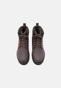 Un paio di stivali in pelle marrone con lacci, dotati di colli imbottiti e suole nere robuste, visti dall'alto su uno sfondo bianco.