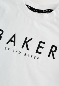 Hvid t-shirt med sort tekst "BAKER BY TED BAKER" trykt over brystet og et sort mærke inden i kraven.
