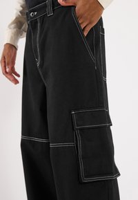 Black cargo pants featuring white stitching, a button closure, side pockets, and a cargo pocket on the thigh. Made from sturdy fabric.
