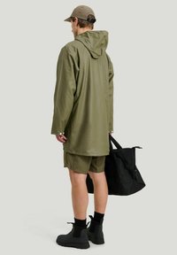 Olivgrön vattentät regnjacka med huva, kombinerad med matchande shorts. Svart väska och svarta ankelskor synliga. Matt, slät yta.